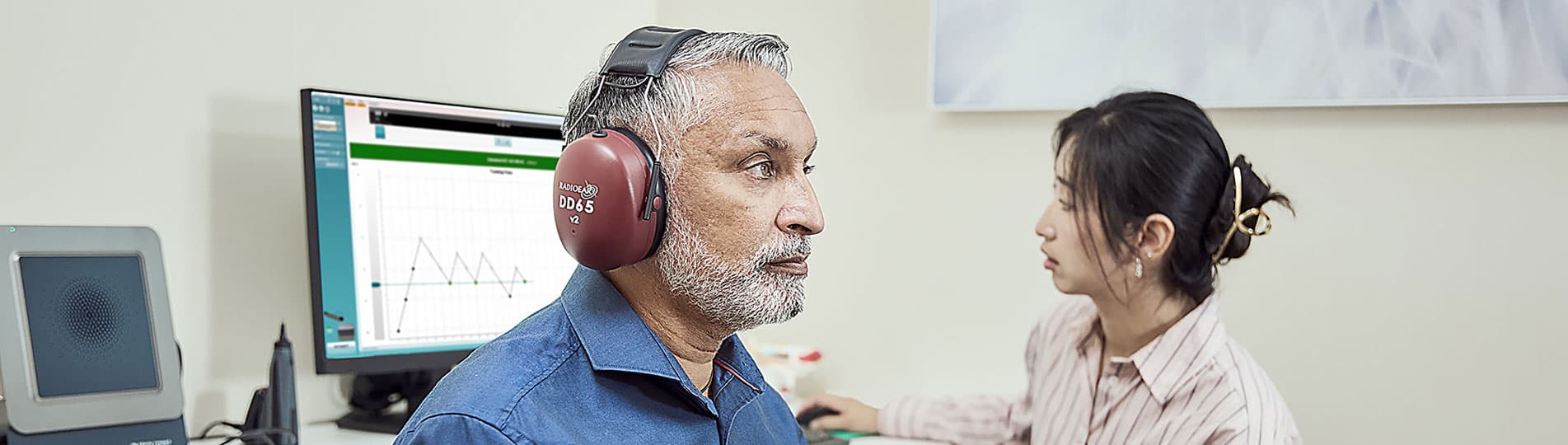 Clinician performing ACT test in client. The client is wearing a blue shirt, has headphones on, and is holding a patient response button. In the background, the clinician is operating the ACT software on a computer screen.