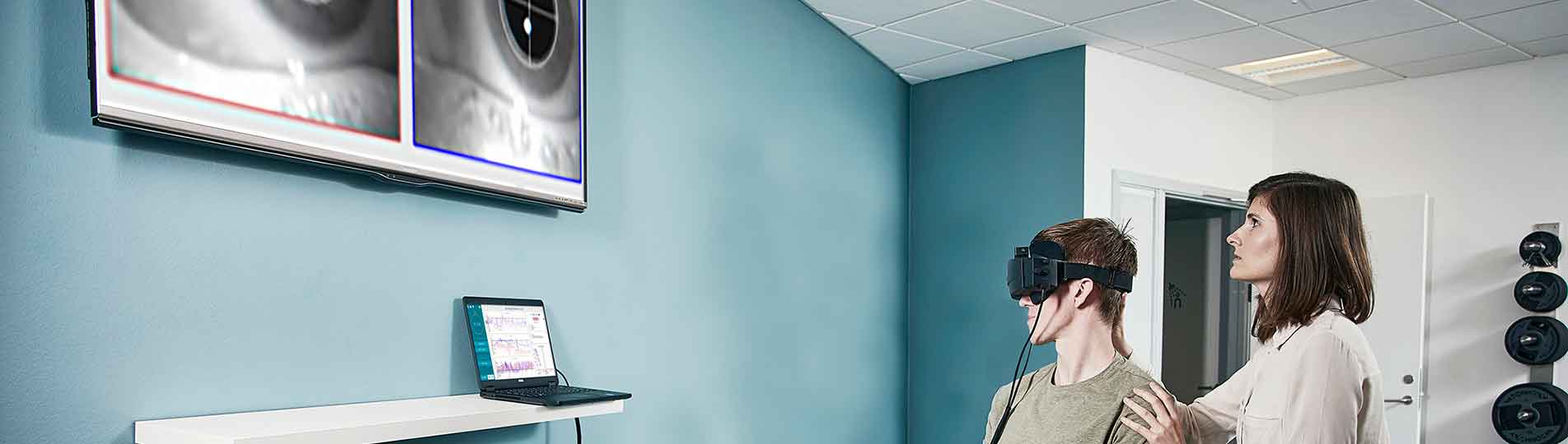 Female clinician performing Advanced Dix-Hallpike maneuver in a young man, who is sat on an examination bed situated in a physical therapy clinic. The man is wearing Video Frenzel goggles with a head sensor mounted. The man is looking toward a computer screen displaying Advanced Dix-Hallpike software. The clinician is looking up toward a large TV screen showing a video image of the patient's eyes.