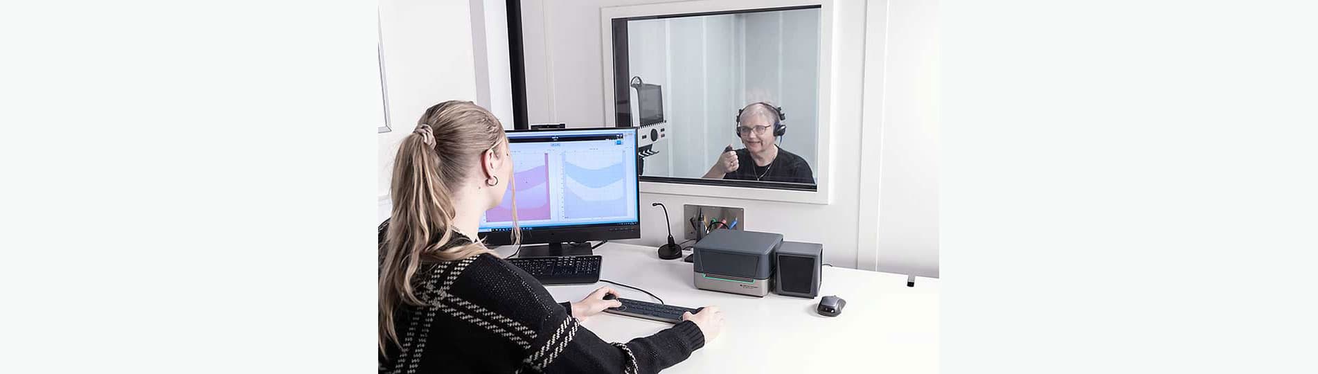 Female clinician sat at a white desk with the Audiometer Keyboard, Affinity Compact, mouse, speaker, microphone, PC keyboard, and a computer screen displaying audiometry software. The clinician is operating the Audiometer Keyboard, with her hands placed on its two dials. The clinician is looking through a window behind which a female patient is sat in a booth. The patient has headphones on and is pressing a patient response button.