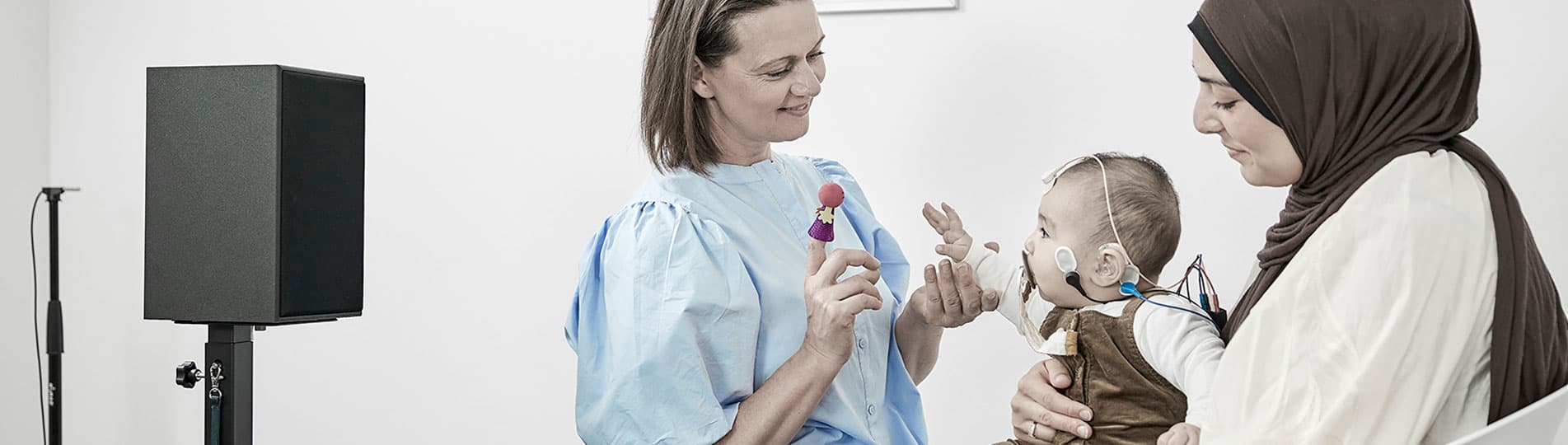 Clinician engaging infant with silent finger puppet while the aided cortical test is running. The infant is sat on their parent's lap, has electrodes placed on their head, and is facing toward the clinician and a speaker.