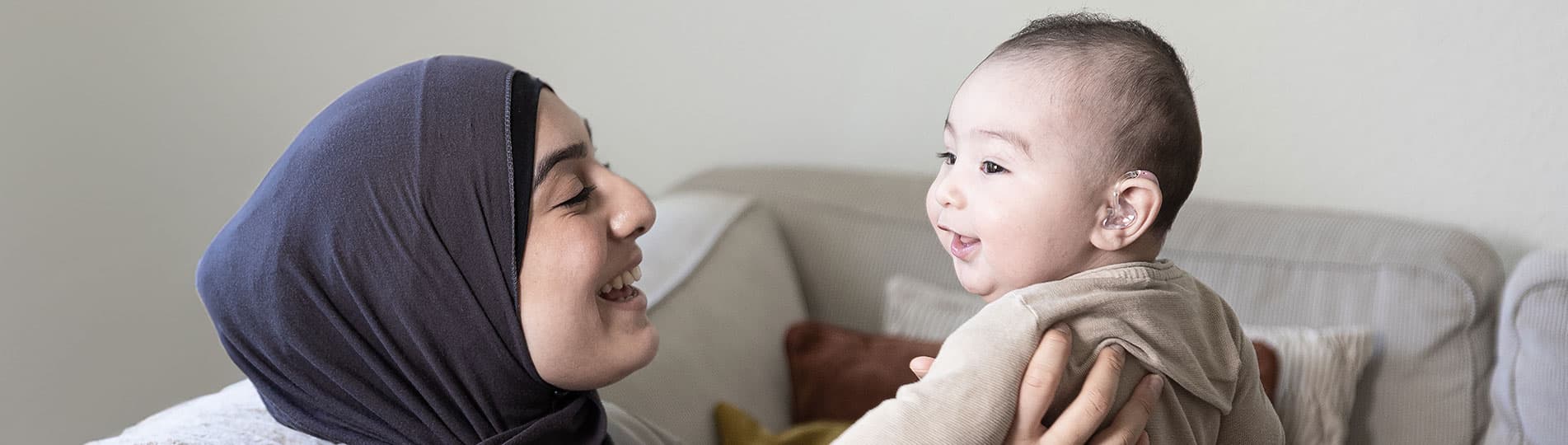 Parent and child enjoying a precious moment on their sofa. The parent is holding the child and the child is smiling and wearing a hearing aid.