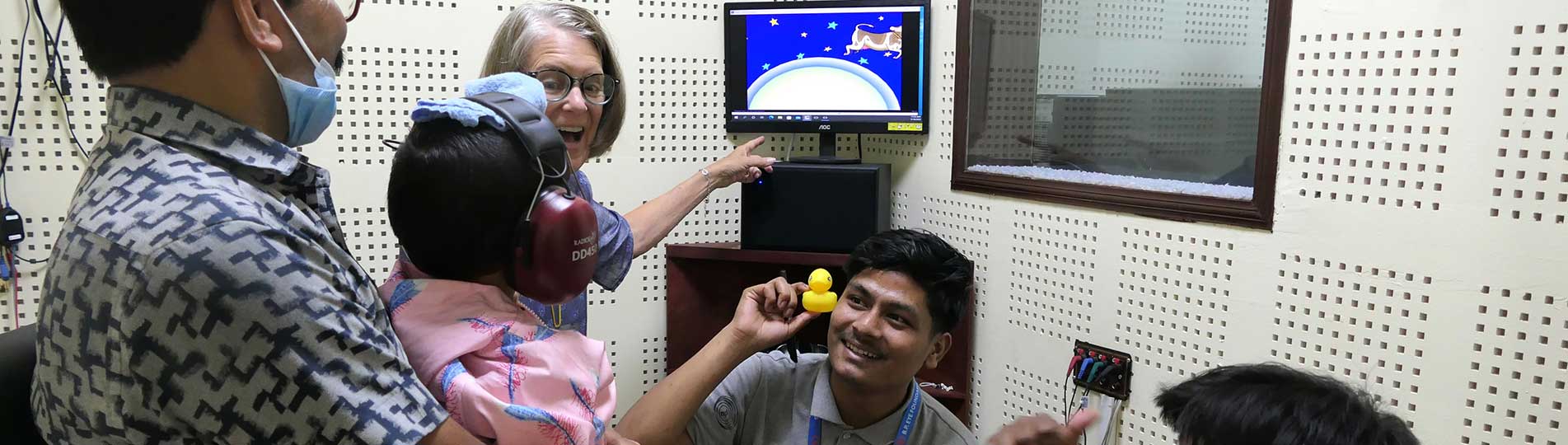 A GFCHL representative and two Nepali CEHWs performing visual reinforcement audiometry in a young child inside an audiometric booth. The child is sat on their father’s lap.