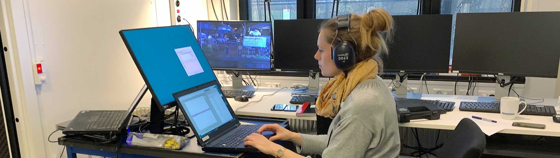 Lisbeth Birkelund Simonsen sat on a chair with wheels and wearing DD65 headphones. Her workstation is full of computer equipment, gadgets, wires, cables and all you could expect for a busy researcher. Lisbeth is working on a laptop, with windows and a view outside to nearby trees to the right of her.