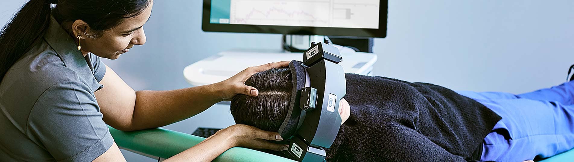 Clinician performing positioning maneuver on patient that is wearing VNG goggles and in a supine position on an examination bed. Computer screen in background showing live eye video and eye movement data